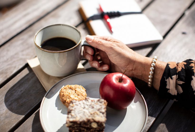 En hand som håller i en kaffekopp och ett fat med frukt och fika. 