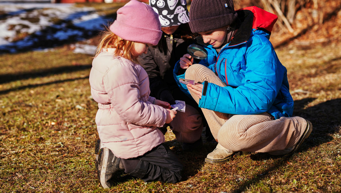 Barn utomhus i varma kläder undersöker en sedel med ett förstoringsglas