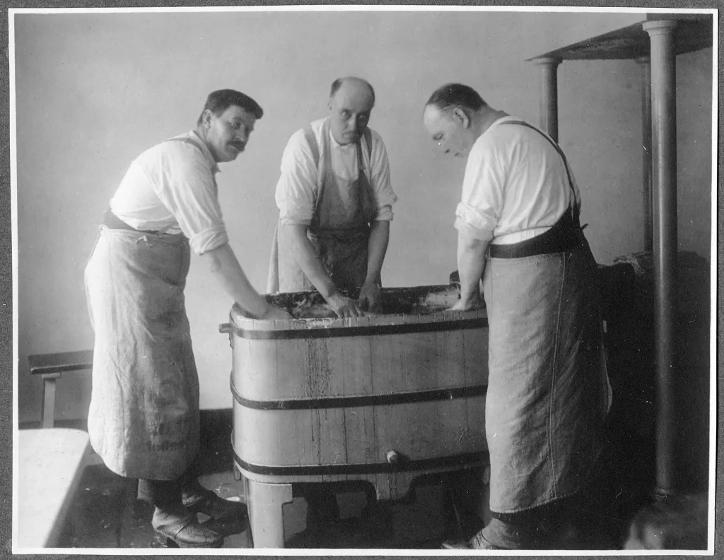 Tre män står vid en trätunna och knodar pappersmassa